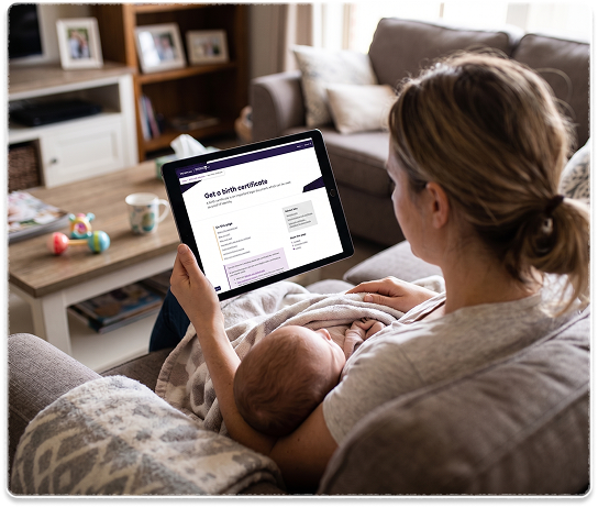 A parent sitting on a sofa holding a baby while using the redesigned “Get a birth certificate” page on a tablet.